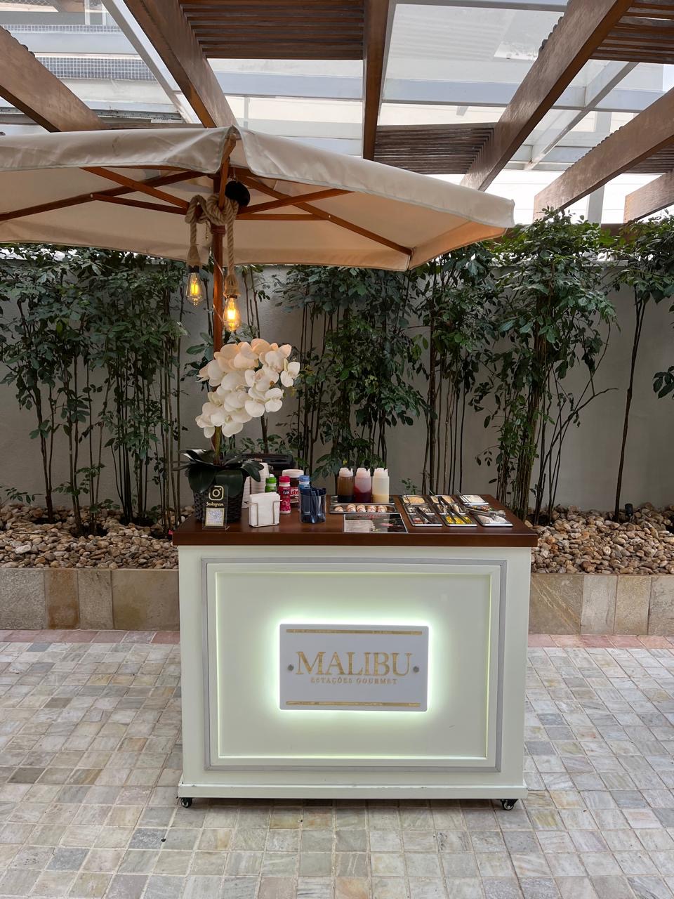 Carrinho gourmet da Malibu Estações Gourmet com letreiro iluminado, ombrelone e orquídeas em área externa